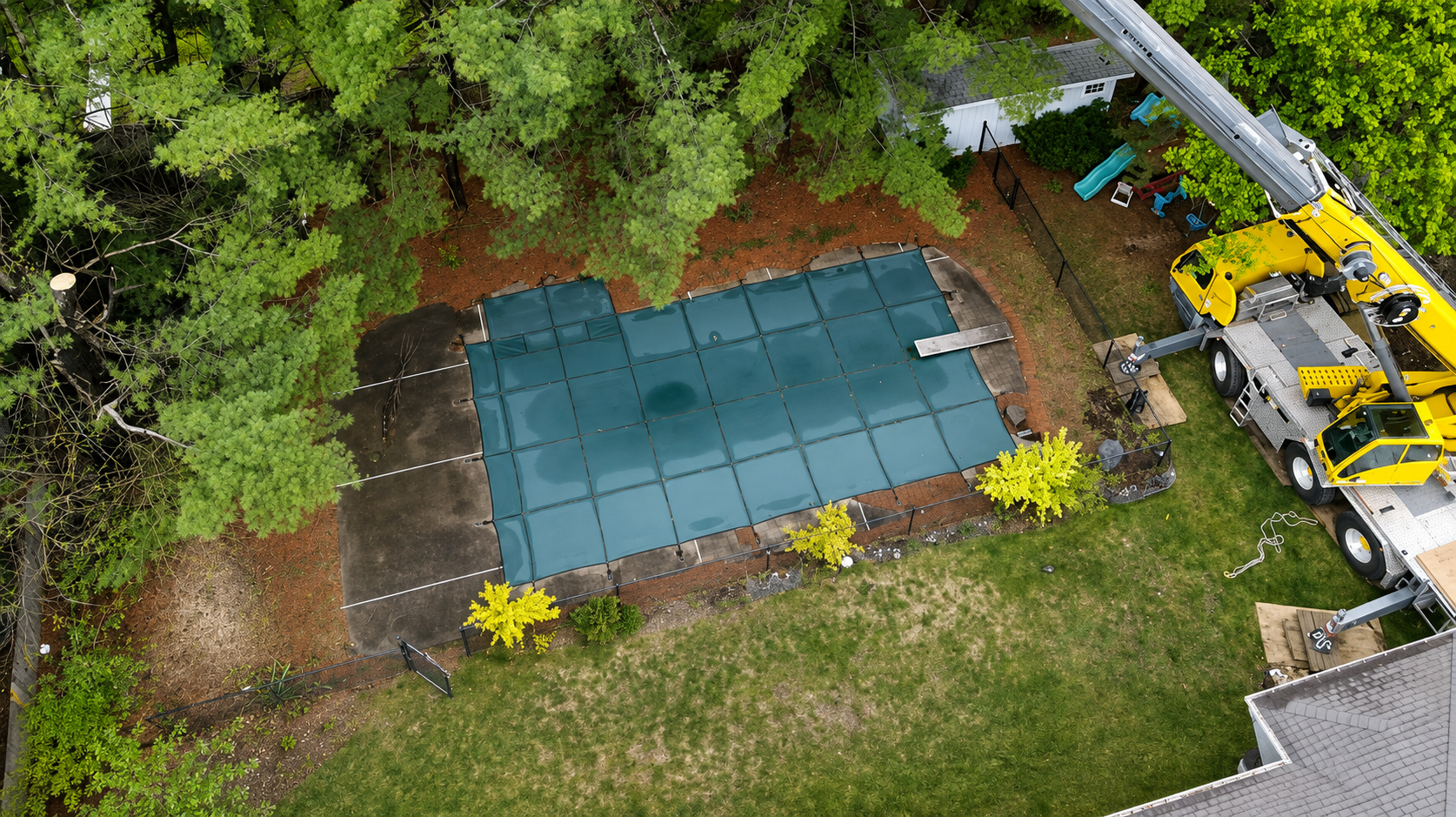 Aerial view of an AA Tree Service crane reaching over a backyard pool to remove trees in a tight-access yard